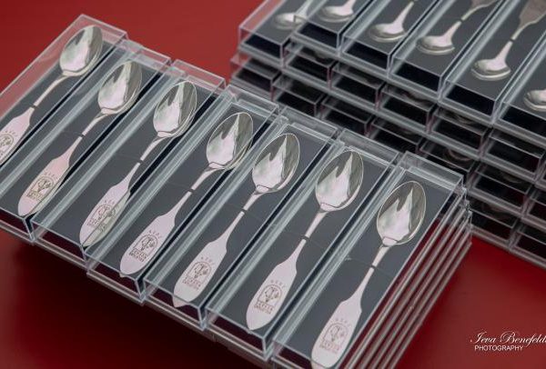 Iesaiņotas sudraba karotes, sakrautas rindās un kaudzēs uz sarkana fona. Katra karote atsevišķā caurspīdīgā plastmasas kastītē ar melnu ieliktni. Uz to rokturiem iegravēts brieža galvas simbols un teksts "SVEICAM VECĀKUS".