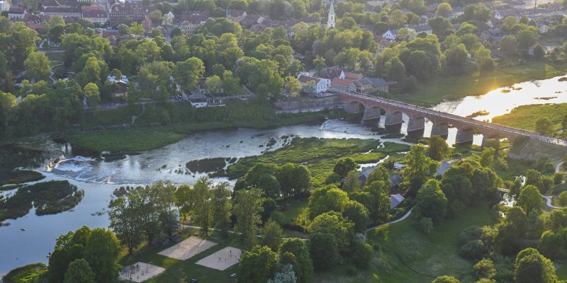 Skats no augšas uz Kuldīgas veco ķieģeļu tiltu, kas šķērso Ventas upi. Upes krastos izvietojusies pilsēta ar ēkām un baznīcas torni, ko ieskauj bagātīgi zaļi koki. Uz upes virsmas labajā pusē atspīd saule, un kreisajā krastā redzami smilšu laukumi un rotaļu laukums.