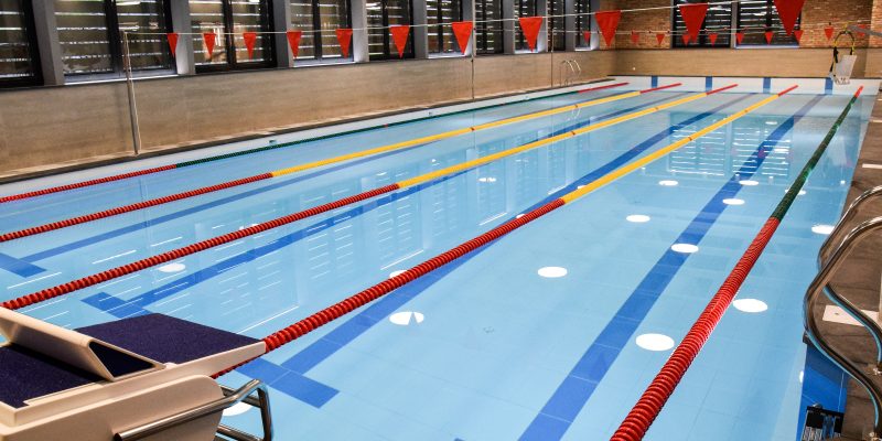 Kuldīgas peldbaseins. Iekštelpu peldbaseins ar gaiši zilu ūdeni, vairākām zilām celiņu līnijām un sarkani dzeltenām celiņu virvēm. Priekšplānā redzams starta bloks ar numuru "1", bet virs baseina izkārtoti sarkani karodziņi.