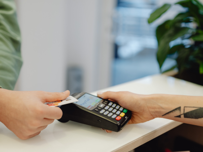 Divas rokas veic maksājumu, ievietojot kredītkarti maksājumu terminālī, ko tur otra roka. Uz termināļa ekrāna redzams uzraksts "PayTel".