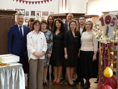 Vienpadsmit cilvēku grupa, trīs vīrieši un astoņas sievietes, pozē fotogrāfijai bibliotēkā. Kuldīgas Galvenās bibliotēkas kolektīvs 105.dzimšanas dienas svinībās.