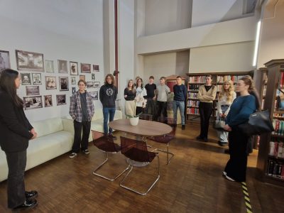 Jauniešu grupa stāv modernā bibliotēkas vai izstāžu zālē, klausoties sievietē, kas stāv priekšplānā pa kreisi. Sienas ir rotātas ar daudzām vecam fotogrāfijām, un labajā pusē ir augsti grāmatu plaukti.