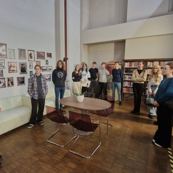 Jauniešu grupa stāv modernā bibliotēkas vai izstāžu zālē, klausoties sievietē, kas stāv priekšplānā pa kreisi. Sienas ir rotātas ar daudzām vecam fotogrāfijām, un labajā pusē ir augsti grāmatu plaukti.