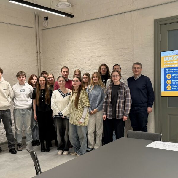 Aptuveni divdesmit jaunieši un viens pieaugušais vīrietis pozē grupas fotogrāfijai gaišā telpā. Aiz viņiem ir liels ekrāns ar informāciju par "JAUNO SPECIĀLISTU SKOLU" un "2. nodarbība – būvvaldes un nekustamo īpašumu speciālisti". Teksts uz ekrāna min: "OTRDIEN 27. janvārī", "No plkst. 13.00 līdz plkst. 17.00", "Vieta: Adatu fabrika", "Mērķauditorija: 8.-12. klases skolēni" un "PIESAKIES ieeja.lv/tilts-s2".