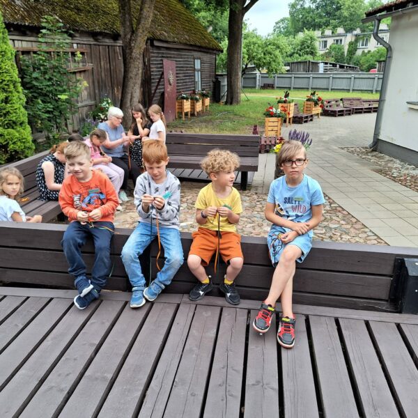 Āra nodarbībā bērnu un pieaugušo grupa veido rokdarbus. Priekšplānā četri zēni sēž uz koka terases, divi no viņiem aktīvi pin krāsainas auklas. Zēnam labajā pusē ir gaiši zils T-krekls ar uzrakstu "SALTY WILD". Fonā redzamas citas personas, vecas koka ēkas un koki.