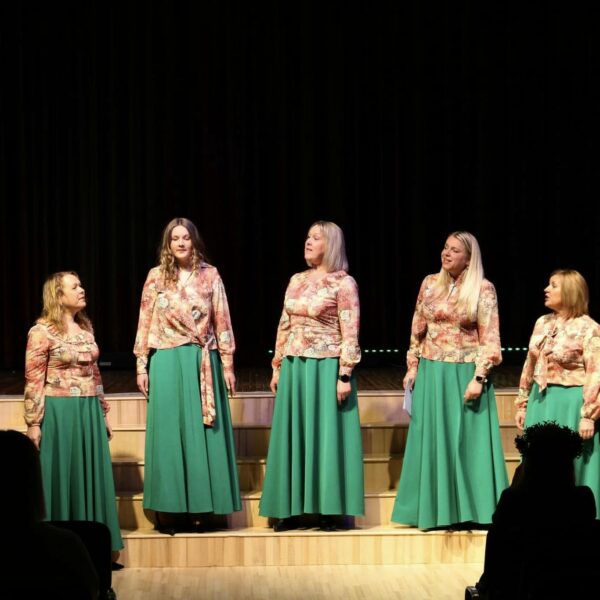 Septiņas sievietes saskaņotos tērpos – ziedu raksta blūzēs un garos, zaļos svārkos – uzstājas uz skatuves. Viņas stāv rindā pret tumšu fonu, dažas skatās viena uz otru, dažas uz priekšu, un viena sieviete dzied ar atvērtu muti. Priekšplānā redzami skatītāju silueti.