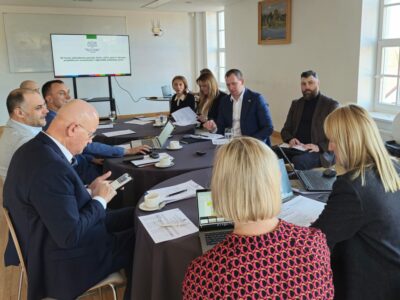 Sanāksme ar aptuveni desmit cilvēkiem, kas sēž pie liela galda, daži strādā ar klēpjdatoriem un pārskata dokumentus. Liels ekrāns rāda tekstu "ES fondu plānošanas periods 2028-2034 gadam Latvijā: priekšlikumi investīcijām reģionālās attīstības jomā".