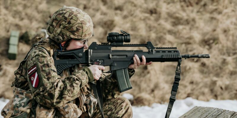 Notupies karavīrs kamuflāžas uniformā ar Latvijas karoga plāksteri un ķiveri mērķē ar šauteni ar optisko tēmekli, valkājot dzirdes aizsargus.