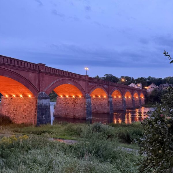 Vecs sarkano ķieģeļu velvju tilts ar akmens balstiem, kas krēslā izgaismots ar silti oranžām gaismām, atspoguļojas upē. Priekšplānā zāļaini krasti.